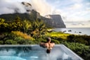 Man relaxing in Capella Lodge infinity pool overlooking Lord Howe Island beach, lagoon, and cloud-capped volcanic peaks at dawn