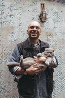 Smiling man Yusuf Çakır holds a grey Scottish Fold cat against a stencil-patterned wall at The Yard Hotel Istanbul.